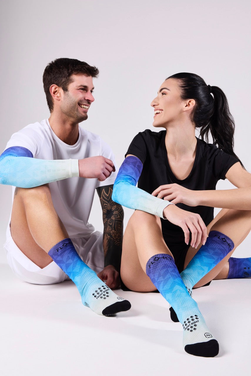 Smiling male and female models sitting together wearing Floky blue ombre arm sleeves and knee-high socks on a grey background, Floky 2025 collection