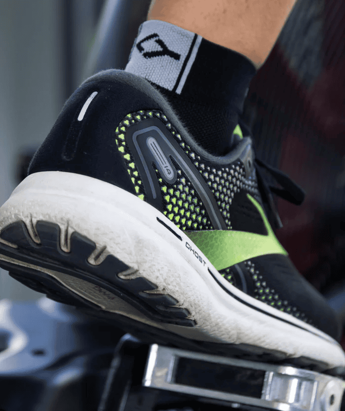 Close-up lifestyle photo of someone on a gym machine wearing a black Floky Multisport short sock with neon yellow Floky logo, paired with a white-soled black running shoe