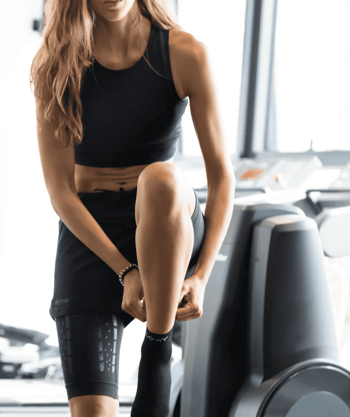 Female athlete in a black sports bra and shorts wearing black Floky Multisport short socks, bending over a gym machine in a bright gym environment