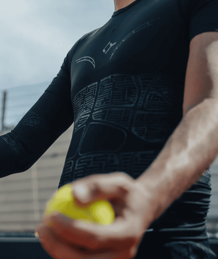 Male athlete wearing black Floky OXY-GEN compression jersey holding yellow padel ball with outstretched arm, showing jersey chest compression panel detailing, outdoor sports court lifestyle action shot