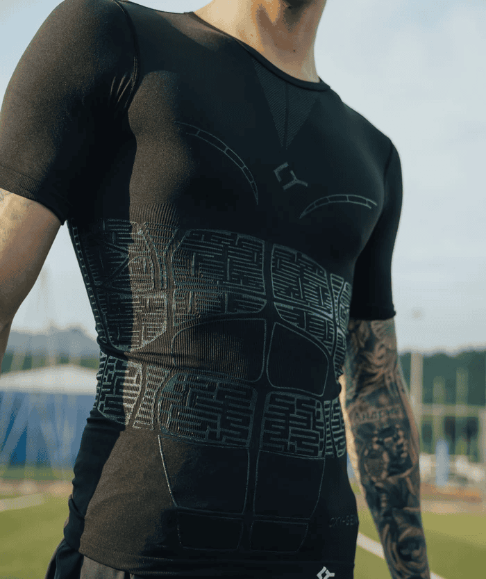 Torso close-up of tattooed male athlete wearing black short-sleeve Floky OXY-GEN compression jersey with teal compression zone grid panel lines, outdoor sports facility lifestyle shot with blue sky background