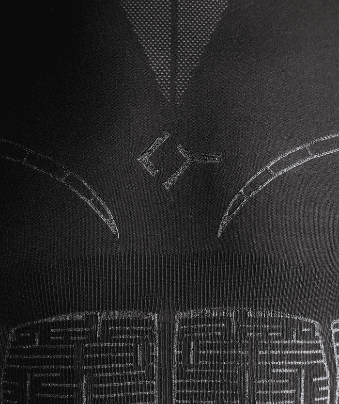 Macro close-up of black Floky OXY-GEN jersey chest area showing V-shaped structural seam lines and geometric compression zone stitching with contrasting dot texture at shoulder, technical fabric detail shot on dark background