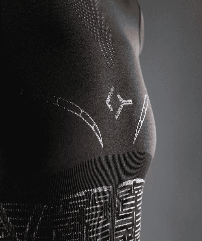 Close-up detail of person wearing black Floky OXY-GEN jersey showing side torso panel with Floky logo in white stitching, curved compression seam lines and geometric grid zones, studio lifestyle detail shot on grey background
