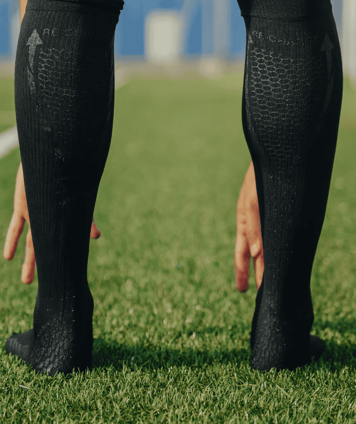 Two athletes standing on green grass sports field wearing black Floky RE-CHARGE over-the-calf compression socks with arrow and textured scale pattern, close-up of legs from knee down, blue sky background outdoor lifestyle shot