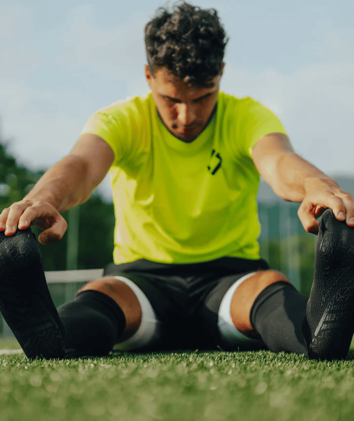 Male athlete sitting cross-legged on grass field stretching forward to touch feet, wearing neon yellow jersey and black shorts with black Floky RE-CHARGE over-the-calf compression socks and white calf support, outdoor sports lifestyle shot