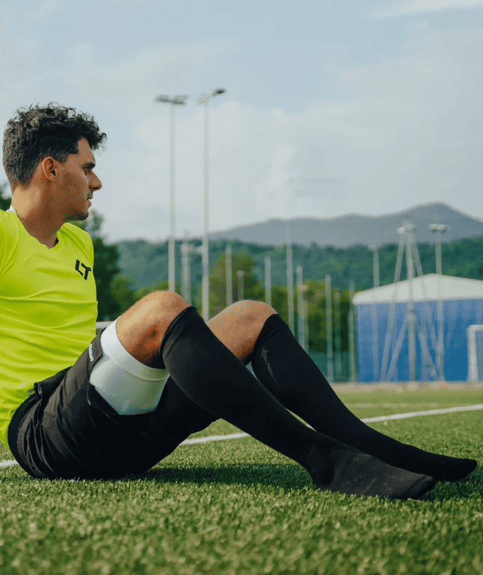 Male athlete sitting on artificial turf sports field wearing neon yellow jersey and black shorts with black Floky RE-CHARGE over-the-calf compression socks and white calf support sleeve, outdoor stadium lifestyle shot with mountains in background