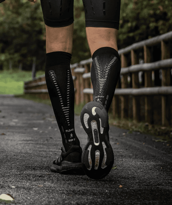 Rear view of runner's legs on forest trail path wearing black Floky RUN UP Long over-the-calf compression socks with vertical stripe detail on calf, black trail running shoes, moody outdoor lifestyle shot with wooden fence and trees in background