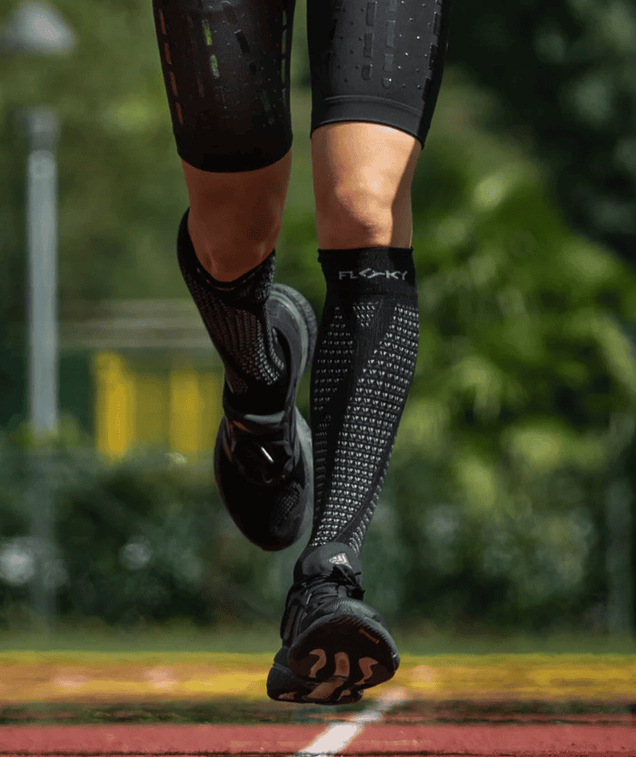 Action shot of runner mid-stride on red athletics track wearing black Floky RUN UP Long over-the-calf compression socks with FLOKY logo, black running shoes raised off ground, dynamic running lifestyle shot with green foliage background