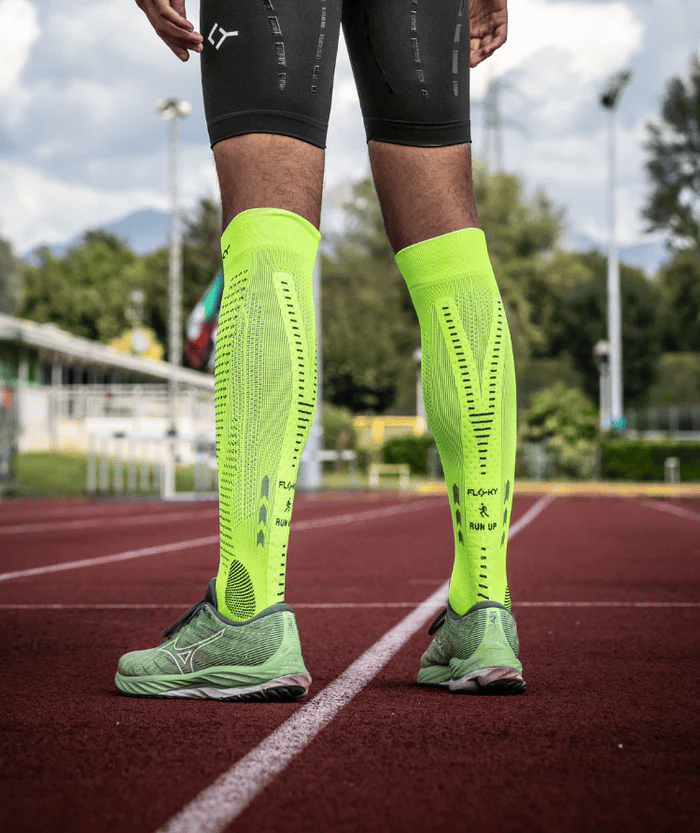 Front view of male legs standing on a running track wearing neon yellow Floky RUN UP Long over-the-calf compression socks with Floky and RUN UP branding visible, outdoor lifestyle shot