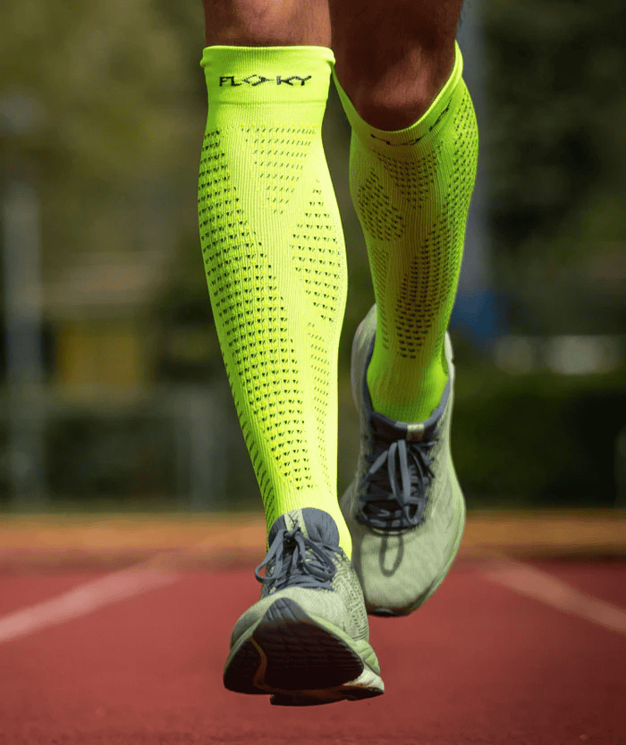 Close-up of runner's lower legs on a red track wearing neon yellow Floky RUN UP Long over-the-calf compression socks and grey running shoes, outdoor lifestyle action shot