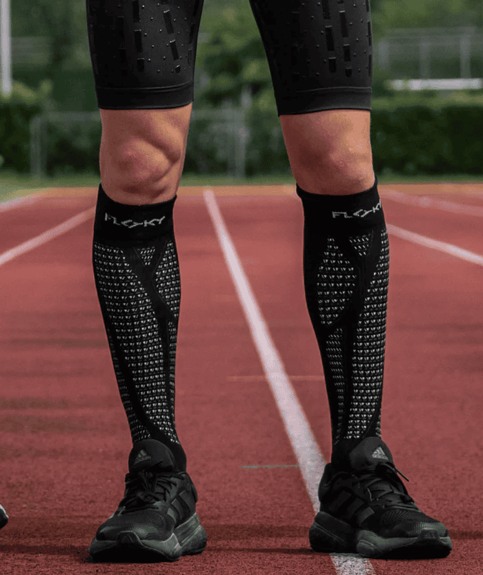 Front view of runner's legs on red athletics track wearing black Floky RUN UP Long over-the-calf compression socks with FLOKY logo on calf, paired with black running shoes, outdoor track lifestyle shot