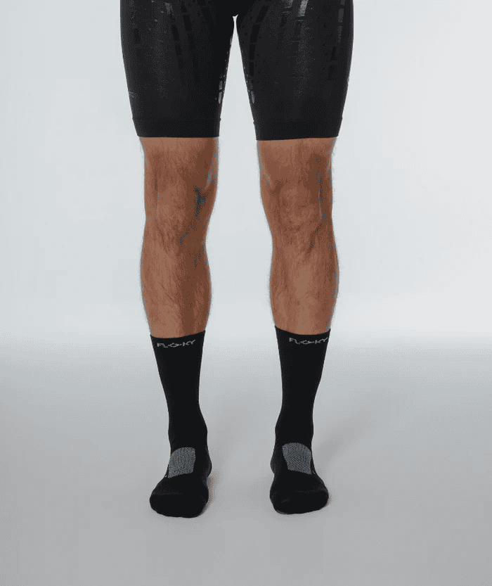 Close-up front view of male legs wearing black Floky RUN UP Medium socks with grey heel detail and black running shoes, studio shot on light grey background