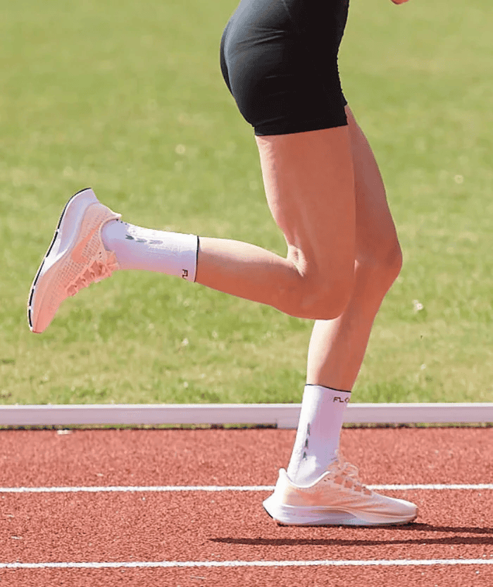Female runner mid-stride on a red running track wearing white Floky RUN UP Medium socks and white running shoes, lifestyle action shot showing lower legs