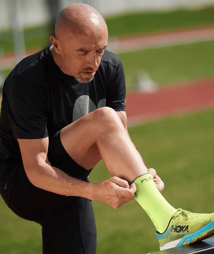 Male athlete putting on a neon yellow Floky RUN UP Medium sock at a running track, close-up lifestyle shot with Hoka running shoe visible