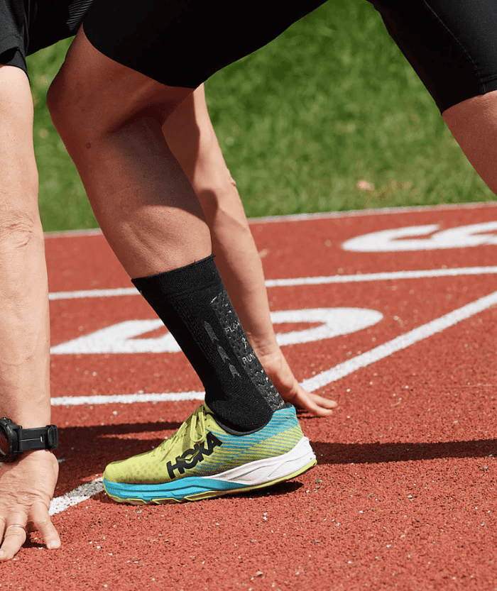 Male athlete in starting position on a red running track wearing a black Floky RUN UP Medium sock with a neon yellow and blue Hoka running shoe, close-up lifestyle shot