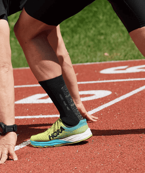 Male athlete in starting position on a red running track wearing a black Floky RUN UP Medium sock with a neon yellow and blue Hoka running shoe, close-up lifestyle shot