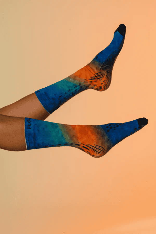 Legs raised showing Floky S-Mash 2.0 socks in blue and orange fusion gradient pattern, lifestyle fashion photo on warm peach background