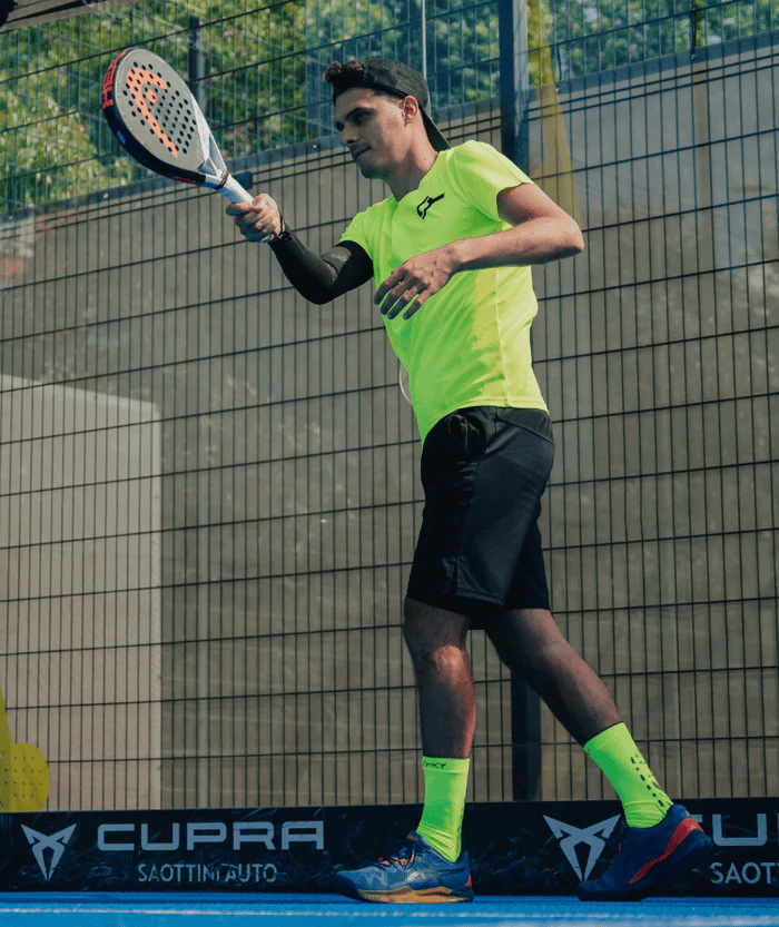 Male padel player in action wearing neon yellow-green Floky S-Mash socks and matching shirt on blue padel court with Cupra branding, lifestyle sports photo