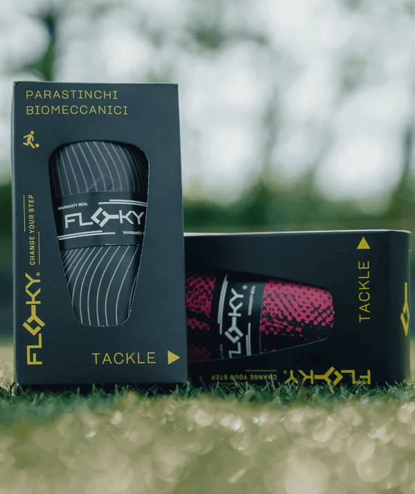 Floky Tackle biomechanical shin guard packaging on grass showing two product boxes with grey wavy and pink shin guard designs, lifestyle product photo