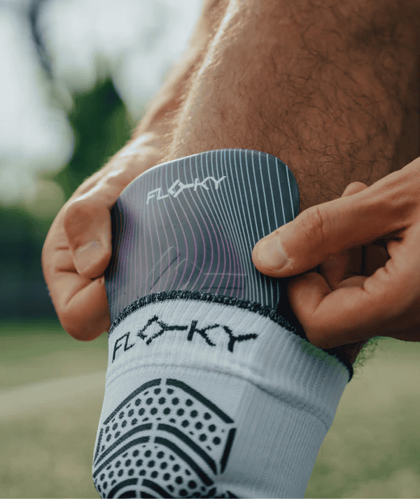 Athlete fitting Floky Tackle shin guard over white grip-dot sock on grass, close-up lifestyle sports photo