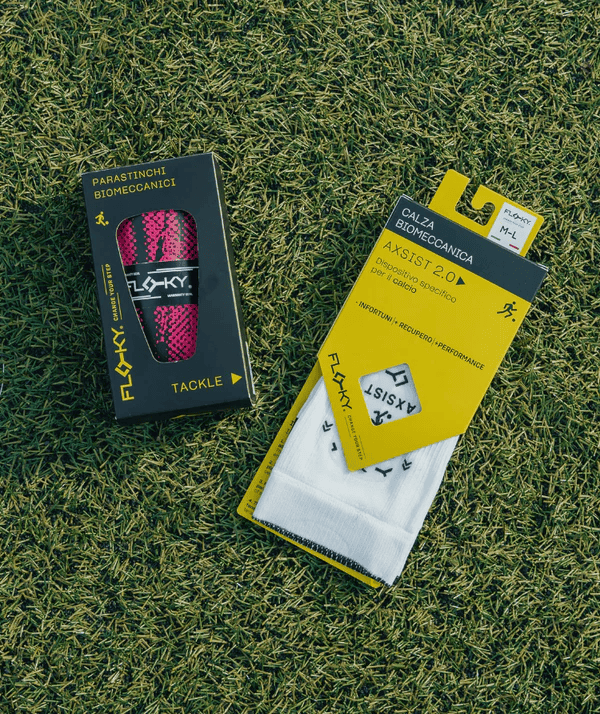 Floky Tackle shin guard packaging on grass showing dark navy box with pink shin guards and yellow biomechanical sock packaging, lifestyle product photo