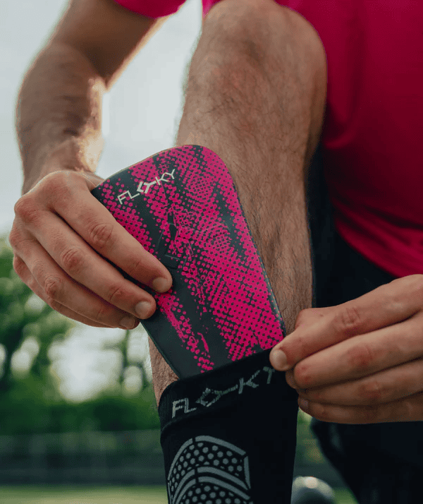 Athlete fitting hot pink Floky Tackle shin guard over white Floky grip-dot sock on leg, close-up lifestyle sports photo