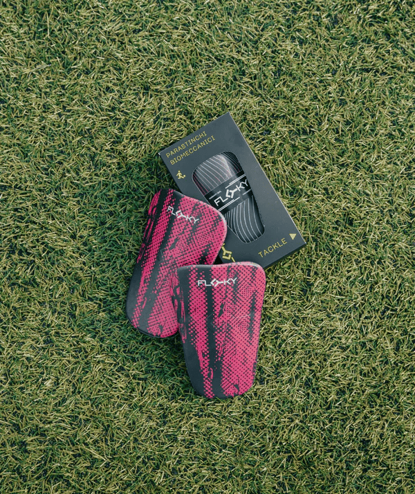 Two Floky Tackle shin guards in hot pink with black Floky logo and dark packaging box on grass, lifestyle product photo