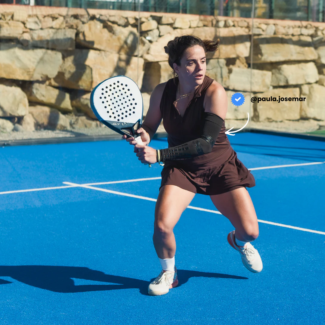 Female padel player Paula Josemar in defensive ready stance wearing black Floky No-Strain arm sleeve on right forearm, dressed in chocolate brown padel dress on blue outdoor court with stone wall background, lifestyle influencer action shot
