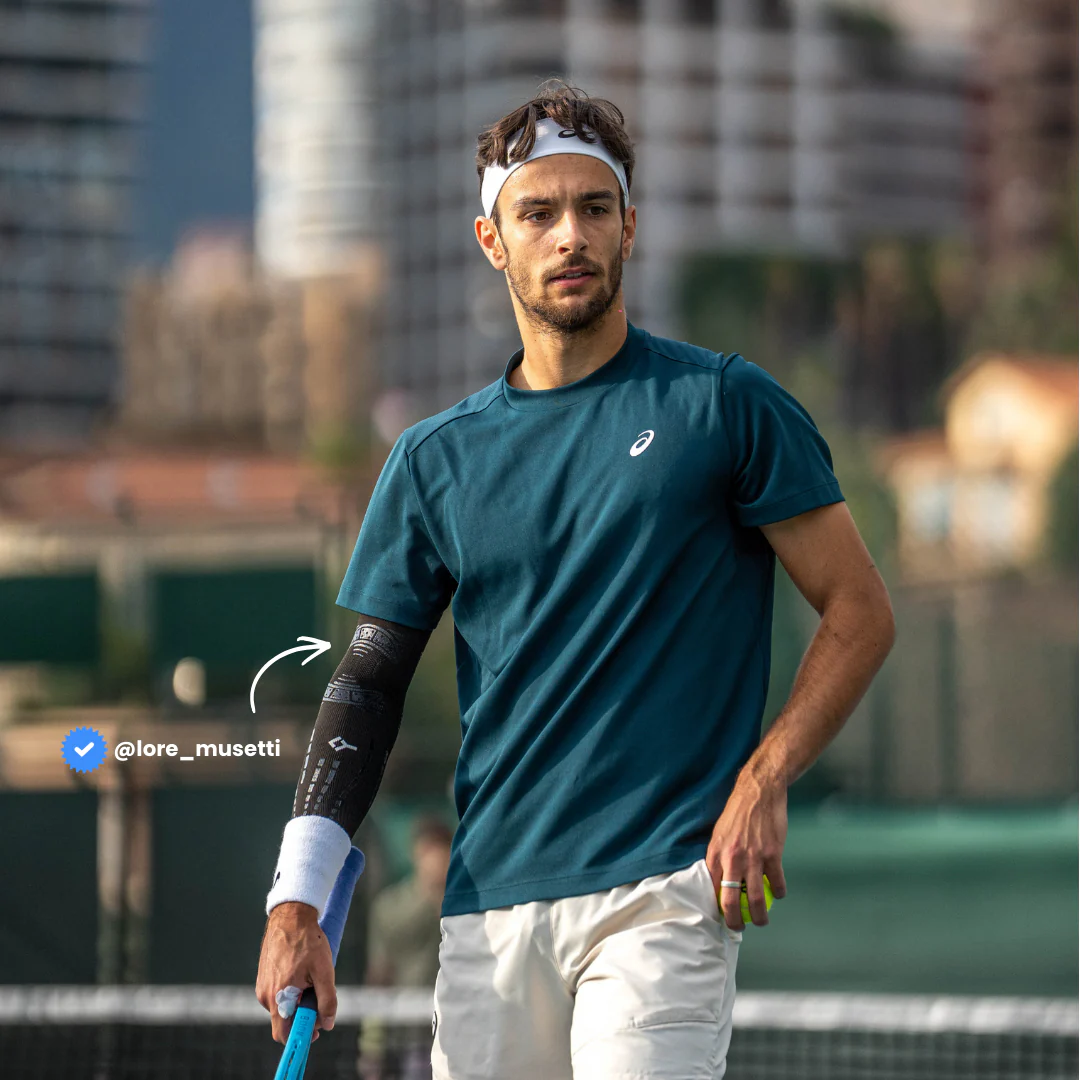 Italian tennis player Lorenzo Musetti wearing black Floky No-Strain arm sleeve on right forearm, dressed in teal Asics shirt and white shorts holding tennis racket on outdoor court with city buildings background, lifestyle influencer shot