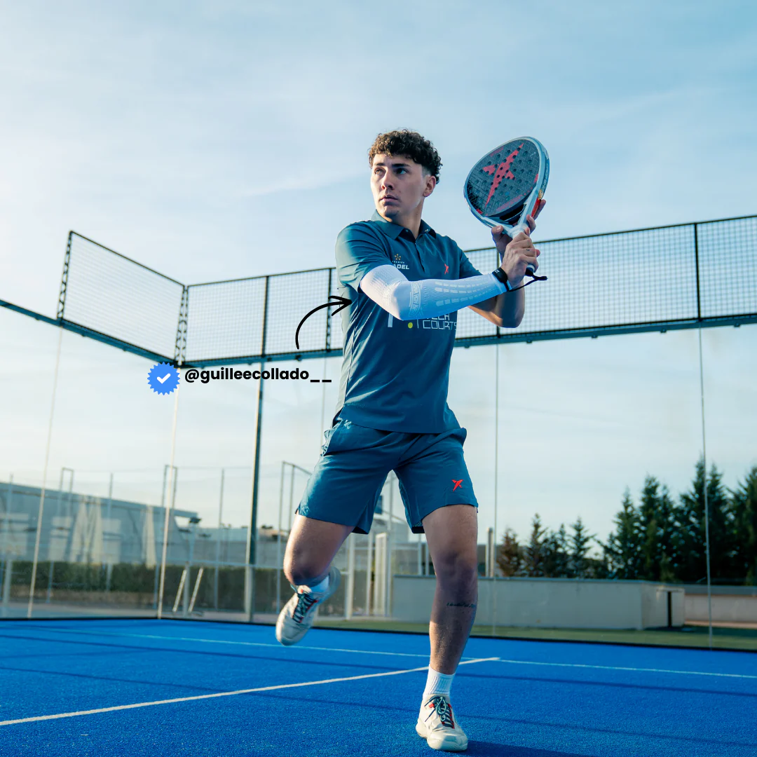 Padel player Guillermo Collado mid-swing wearing white Floky No-Strain arm sleeve on right arm, dressed in navy blue Nox padel kit, low-angle action shot on blue outdoor padel court, lifestyle influencer shot