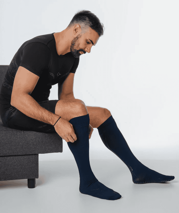 Man sitting on grey couch wearing all-black outfit looking down at his legs wearing navy blue Floky over-the-calf socks, studio lifestyle shot on white-grey background